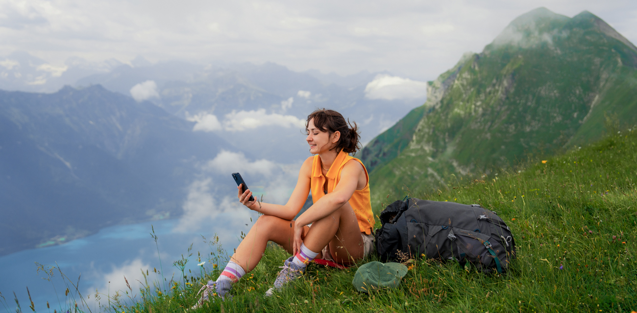 Eine Frau sitzt in sommerlichem Gewand auf einem Berghang und blickt in ihr Smartphone
